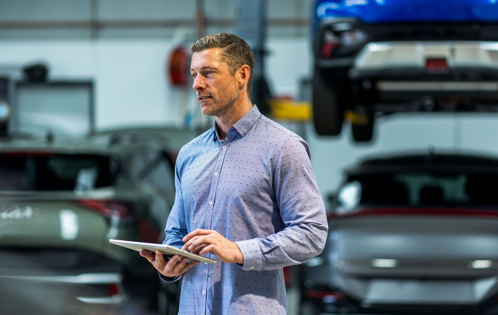 Fleet manager using a tablet in an automotive workshop with cars and a vehicle lift in the background