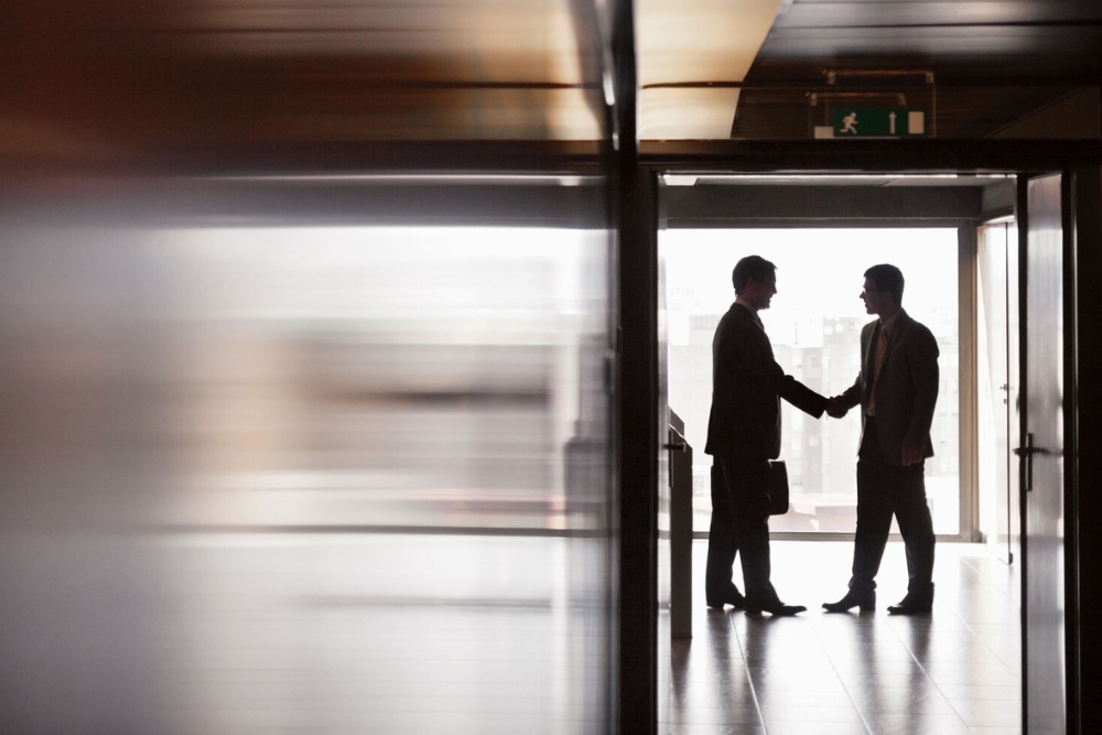 Two business colleagues in suits shake hands in a backlit office corridor, illustrating employee benefits in the automotive sector.