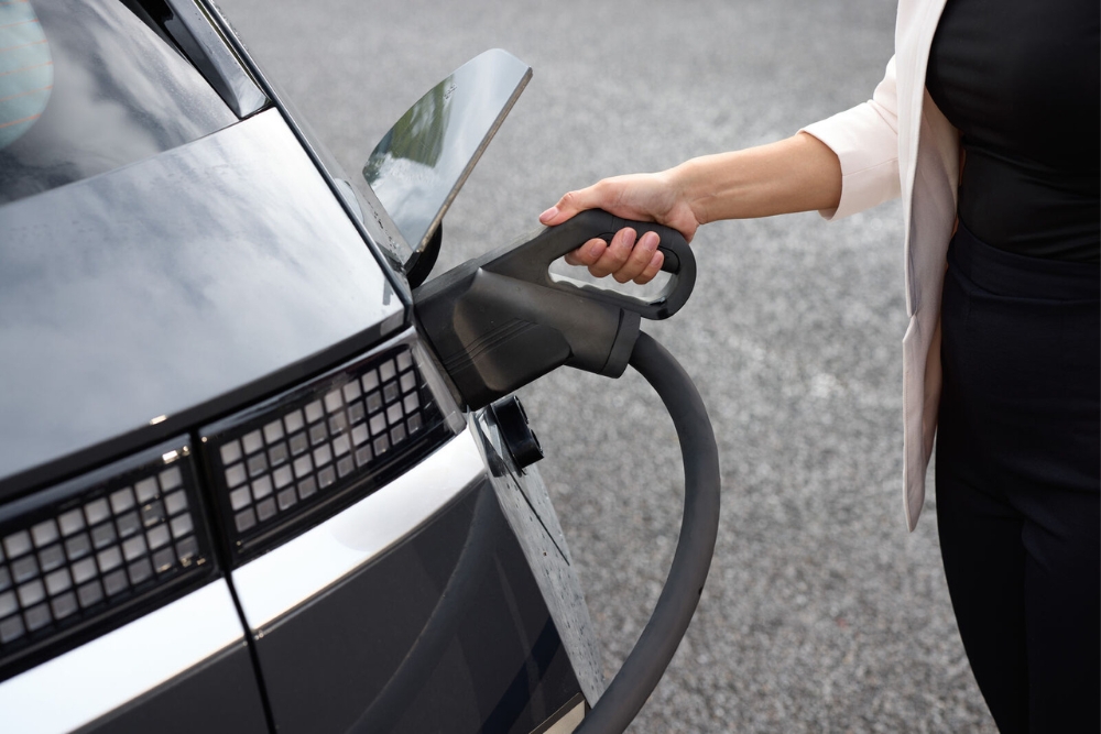 Close-up of a person’s hand inserting a charging plug into an electric car’s charging port on a paved surface, highlighting employee benefits in the automotive sector.