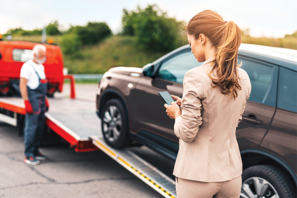 A woman in business attire stands by her parked SUV, looking at her smartphone while a tow-truck operator readies the vehicle on a flatbed, reflecting employee benefits in the automotive sector.
