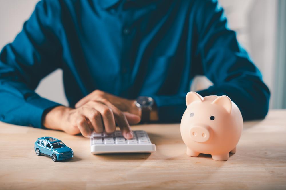 How do I report a company car on a P11D form UK: A man in a blue shirt using a white calculator at a wooden desk, with a small blue toy car on one side and a pink piggy bank on the other.