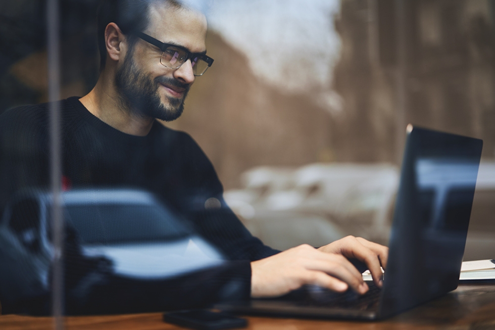 Man smiling while working on a laptop in a café, representing strategic planning or digital management of a growing business fleet.