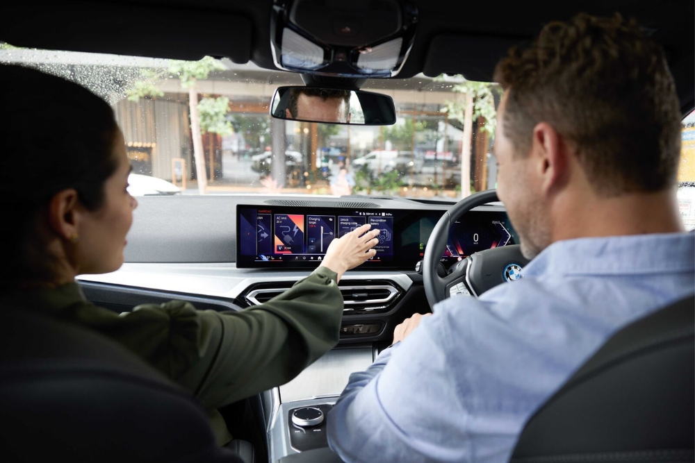 Interior view of a driver and passenger interacting with a wide touchscreen infotainment display on a car dashboard, featuring semiconductor-powered interface display.