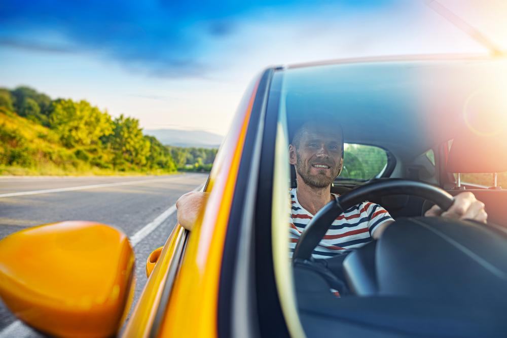 Short-term vehicle hire: A smiling man with a beard driving a yellow car along a country road on a sunny day, viewed through the windscreen with green hills in the background.